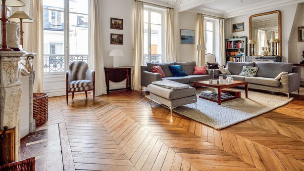 Een ruime woonkamer met een visgraatvloer, een open haard, een gestreepte fauteuil en een groot raam met gordijnen.