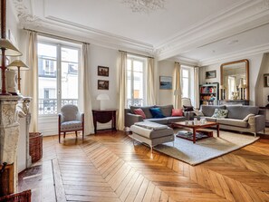 A spacious living room with a herringbone floor, a fireplace, a striped armchair, and a large window with curtains.