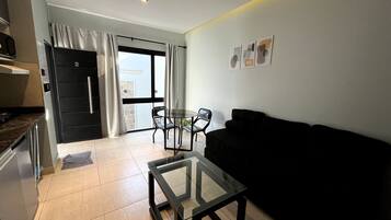 A modern living room with a glass coffee table, a black sofa, and a dining area with chairs.