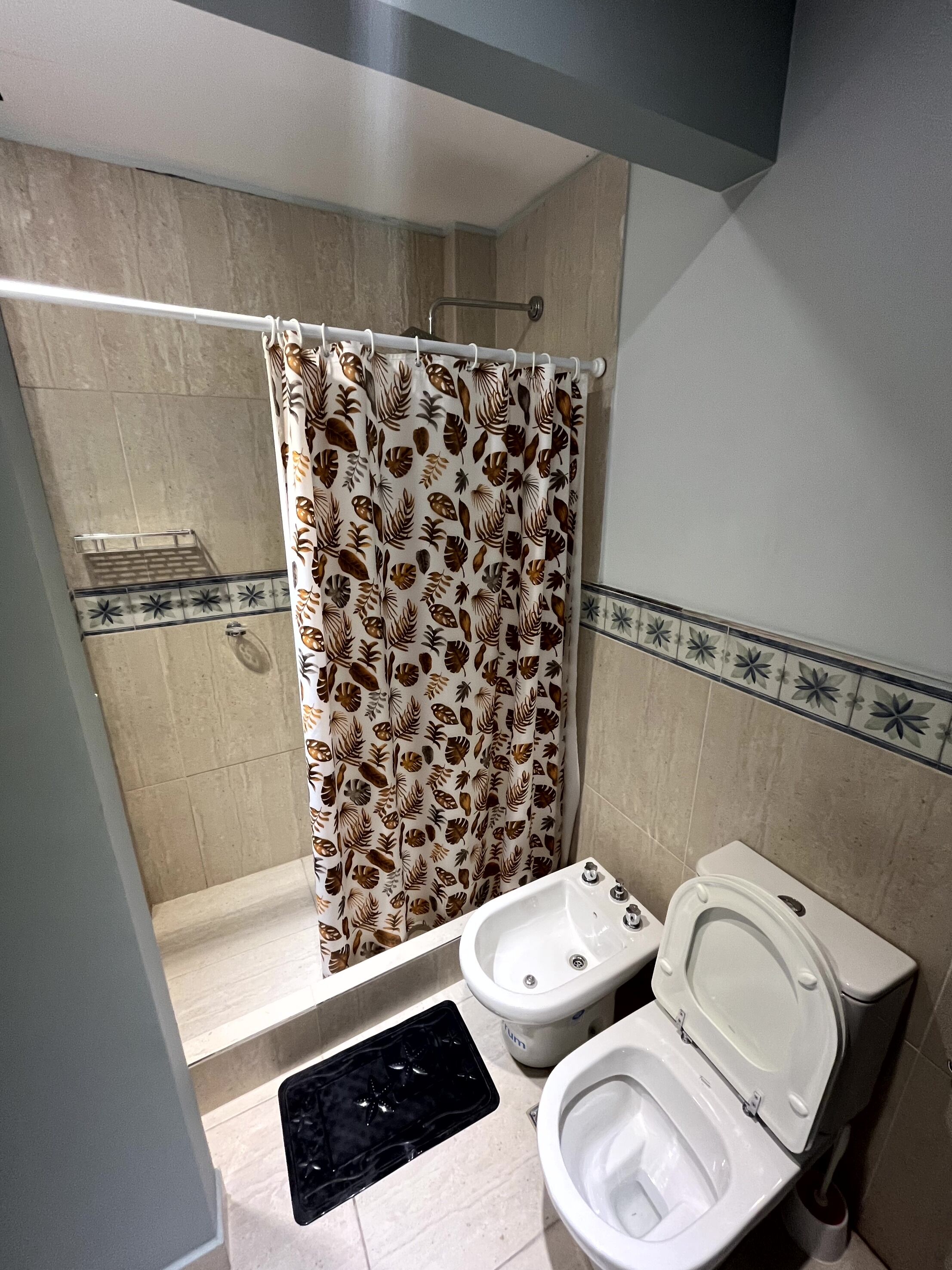 Une salle de bain équipée d’une douche, de toilettes, d’un bidet et d’un tapis noir sur le sol.