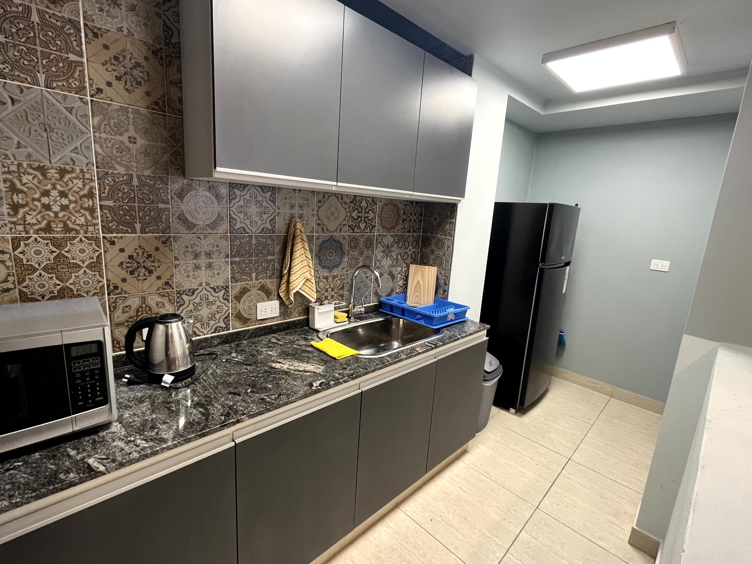 A kitchen with patterned tiles, a microwave, a kettle, a sink with a blue dish rack, and a black refrigerator.
