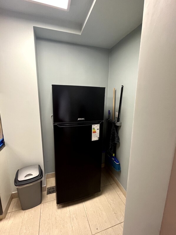 A compact kitchen with a black refrigerator, a trash bin, and a broom leaning against the wall.