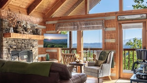 A cozy cabin interior with a stone fireplace, a comfortable leather couch, a wooden deck with a view of the mountains, and a large window with a scenic landscape.