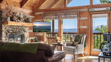 A cozy cabin interior with a stone fireplace, a comfortable sofa, a dining table, and a large window offering a scenic view.