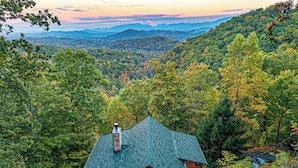 A cabin with a green roof surrounded by dense forest, offering a view of distant mountains.