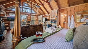 A cozy log cabin interior with a large bed, two glasses of wine on a tray, wooden furnishings, and a ceiling fan.