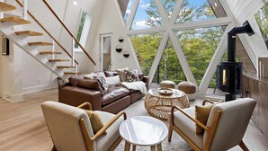 A spacious loft with a high ceiling, a wooden staircase, a cozy seating area with a fireplace, and large windows offering a view of the outdoors. - Sky-High A Frame Retreat Hottub & EV charging (Beech Mountain)