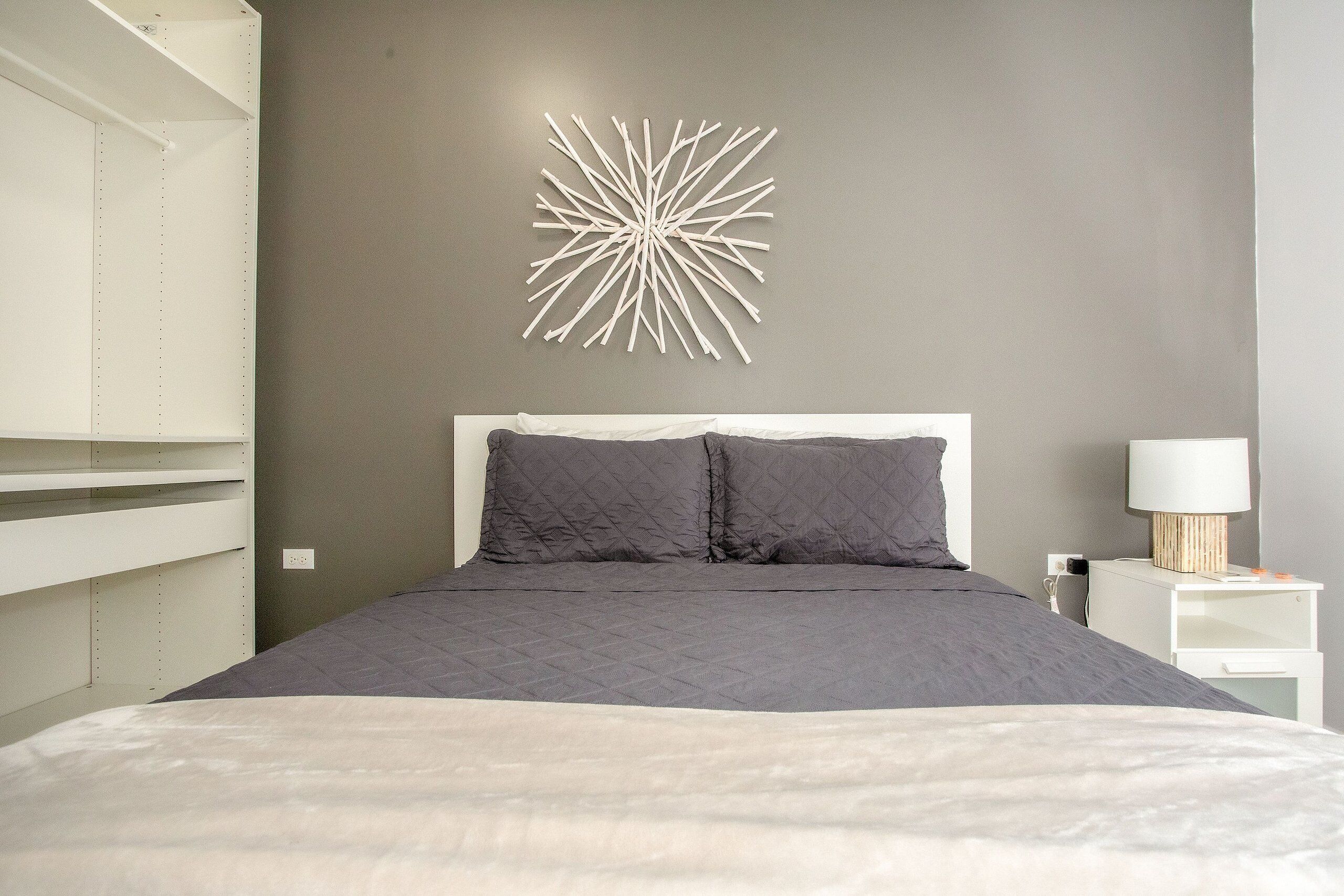 Un dormitorio con cama, ropa de cama gris, un aplique en la pared y una mesita de noche con lámpara.