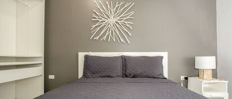 Un dormitorio con cama, ropa de cama gris, un aplique en la pared y una mesita de noche con lámpara.