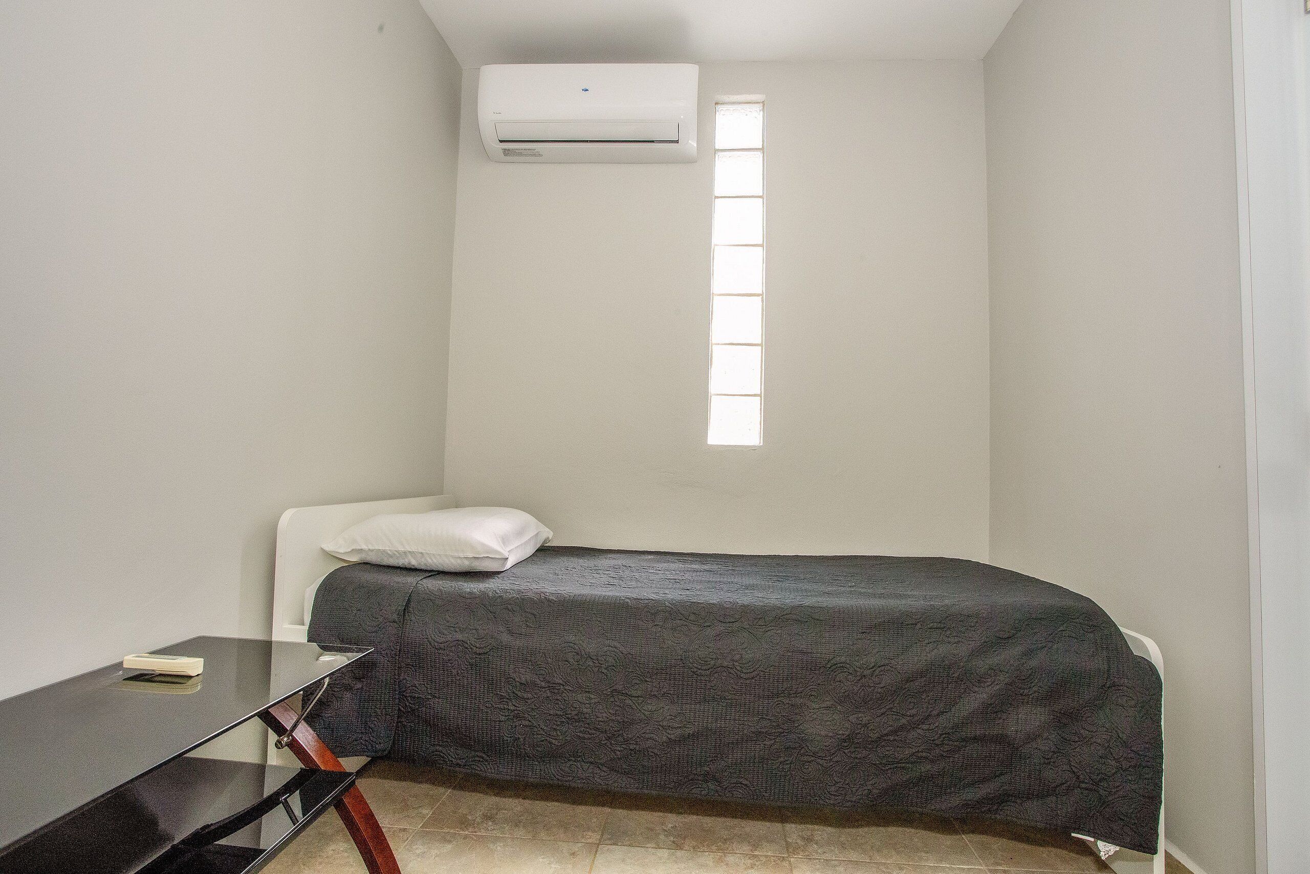 Una habitación pequeña con cama individual, ventana, escritorio y unidad de aire acondicionado.