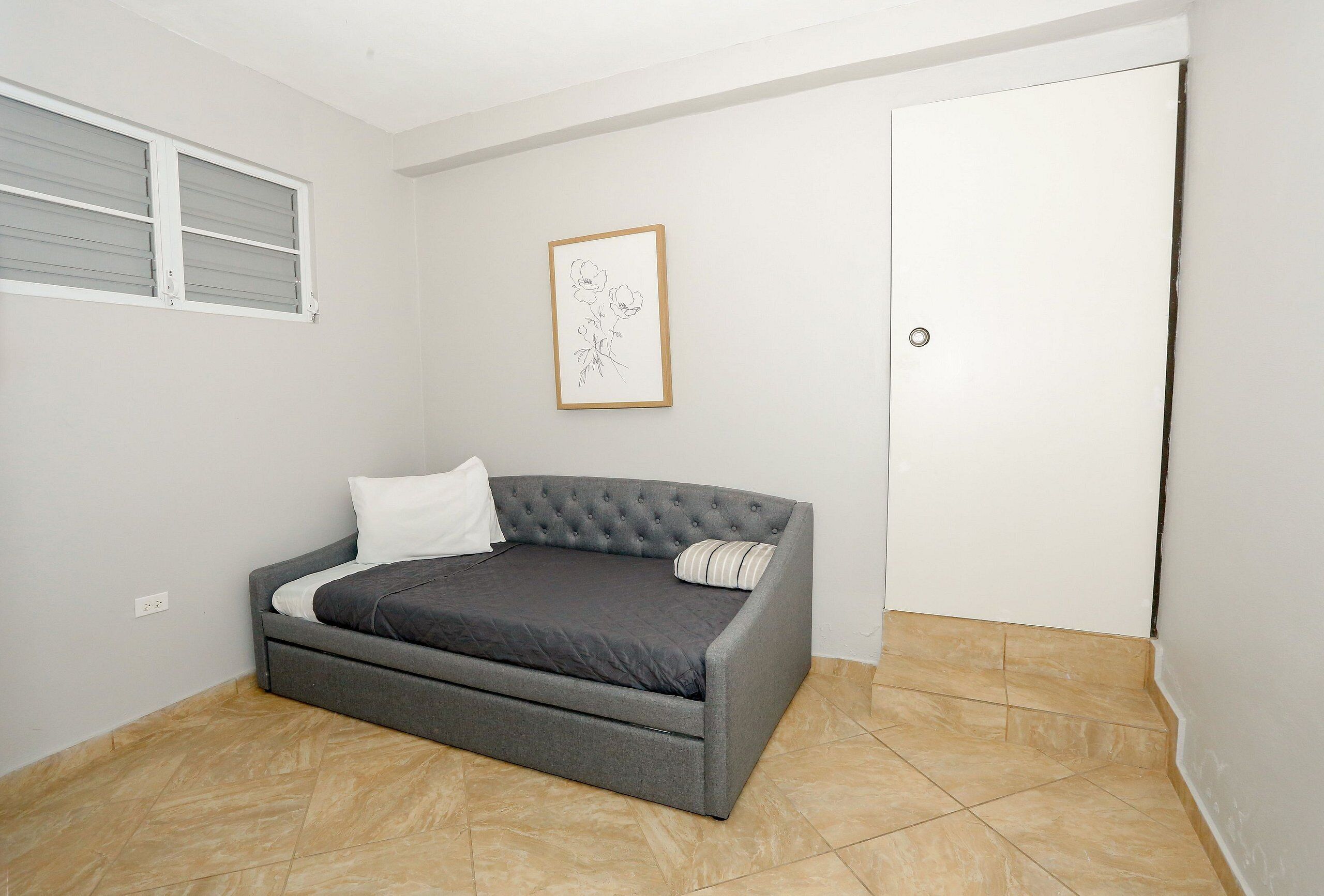 Quarto com um sofá-cama cinzento, uma porta branca e um desenho emoldurado na parede.
