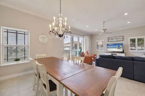 A dining area with a wooden table and white chairs, a chandelier, and a large window with blinds.