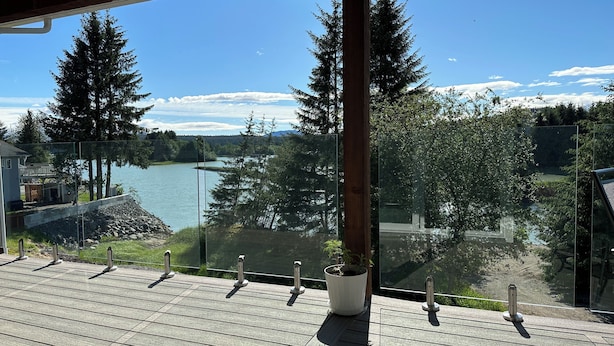 Una terraza de madera con barandilla de vidrio da vista a un lago, rodeado de árboles y una casa.