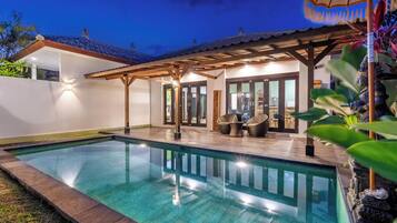 A swimming pool with a wooden deck and outdoor seating, surrounded by a white wall and tropical plants.
