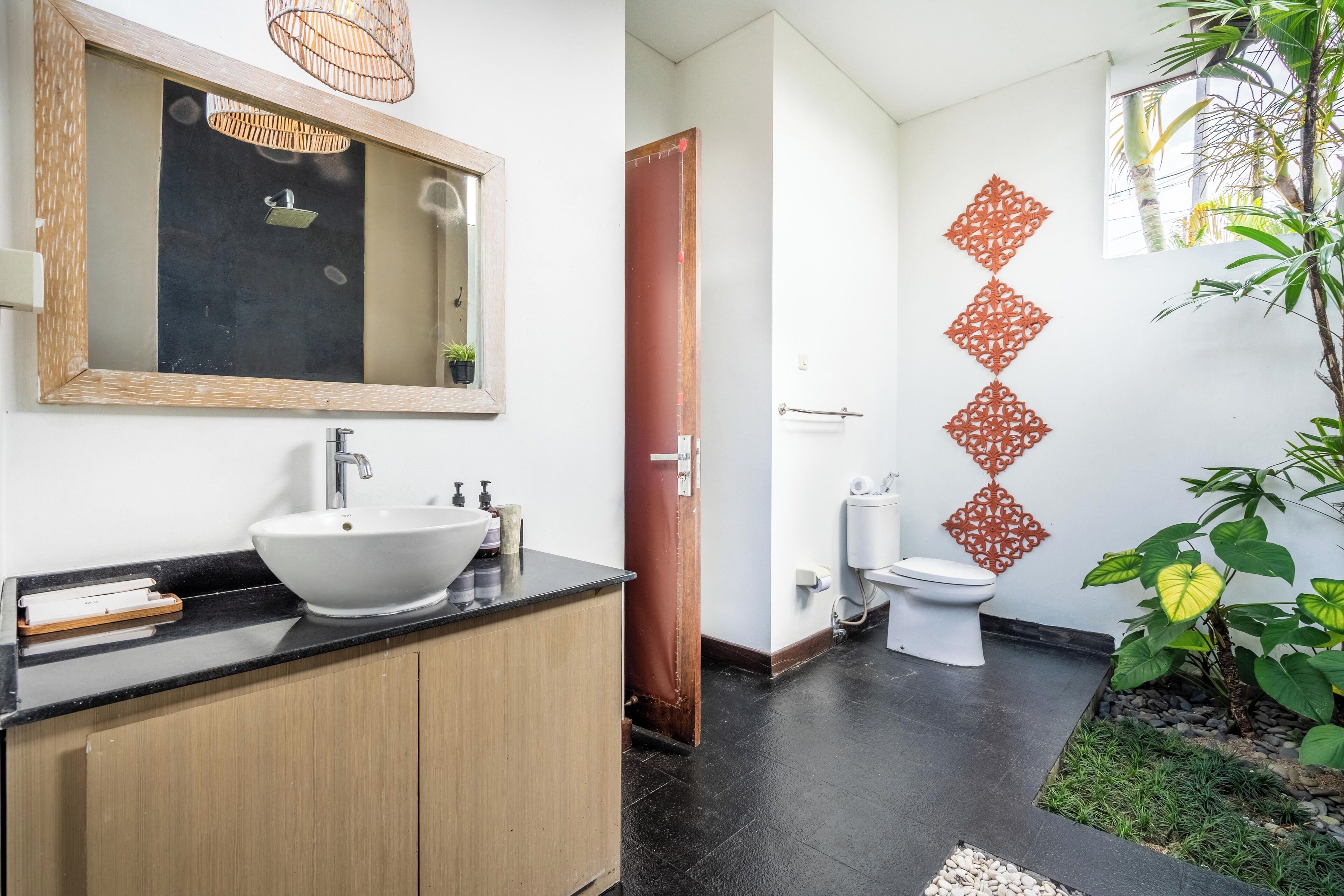 A modern bathroom with a large mirror, a white sink, and a toilet. There is a small plant and decorative wall art.