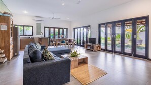 A spacious living area with a sofa, coffee table, and a TV. There is a kitchen area with a dining table and chairs. The room has large windows and a sliding glass door leading to an outdoor area. - 888 Residences - Complex of Private Villas with Pools (Ubud)