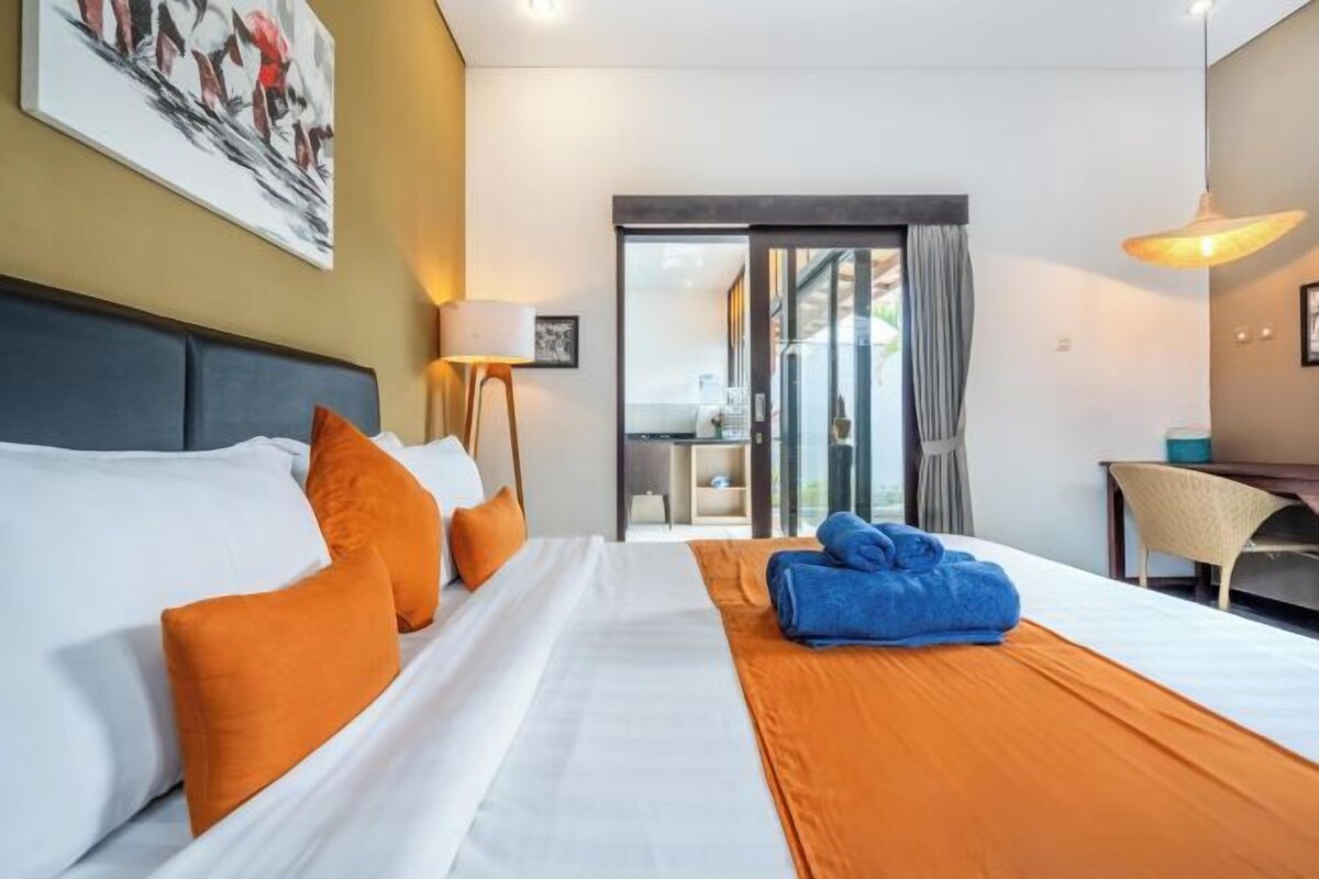 A hotel room with a bed, orange and white bedding, a blue towel, a desk, a chair, and a painting on the wall.