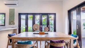 A dining area with a wooden table and chairs, a decorative wall piece, and a view of greenery through a glass door. - 888 Residences - Complex of Private Villas with Pools (Ubud)