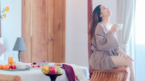 Eine Person sitzt auf einem Holzstuhl am Fenster und hält eine Tasse. Ein Frühstückstablett mit Früchten und einem Glas Orangensaft steht auf dem Bett.