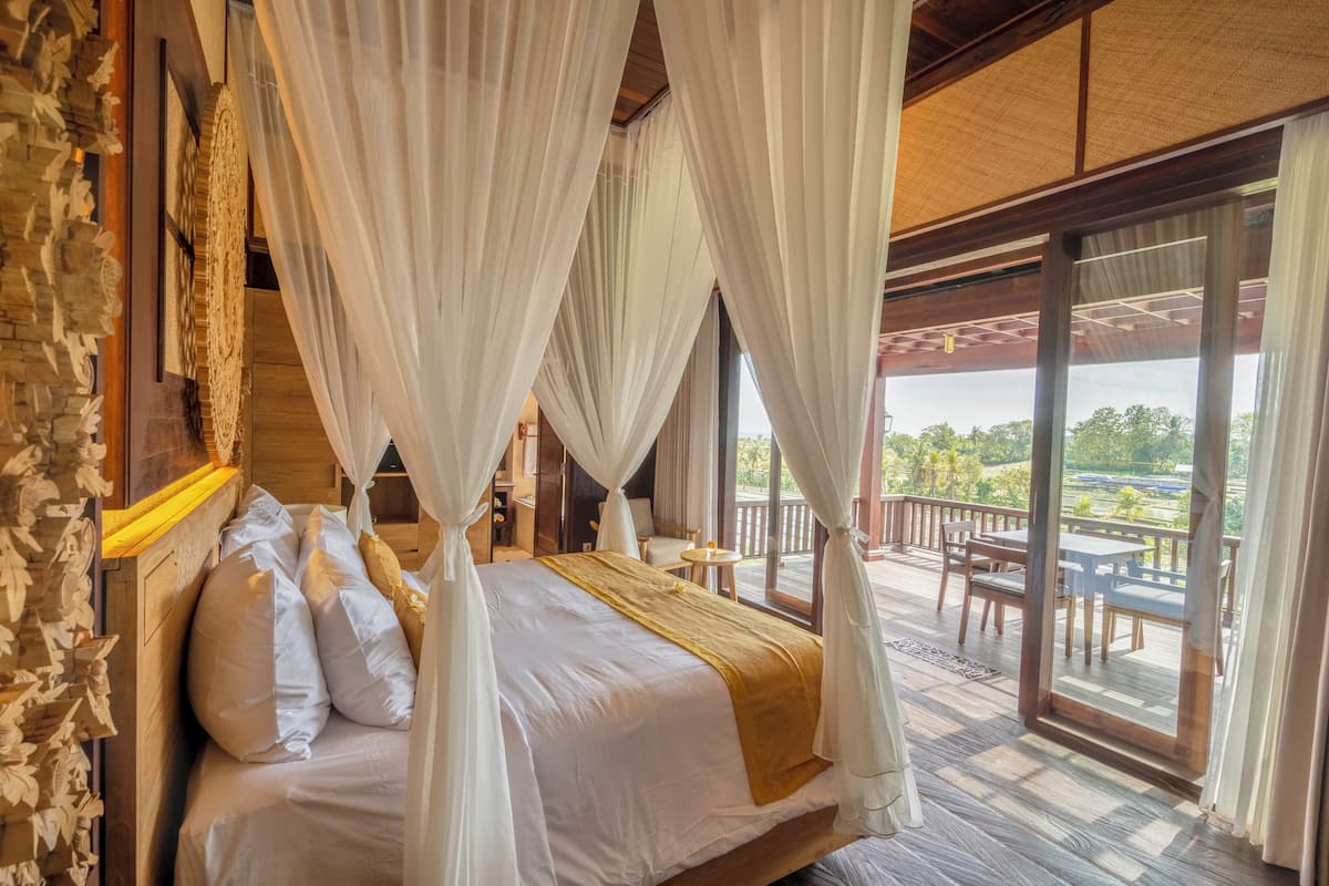 Una habitación con cama, cortinas blancas, cabecero de madera y un balcón con mobiliario exterior.