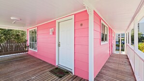 Un bâtiment rose avec une porte et des fenêtres blanches, une terrasse en bois et une balustrade blanche.