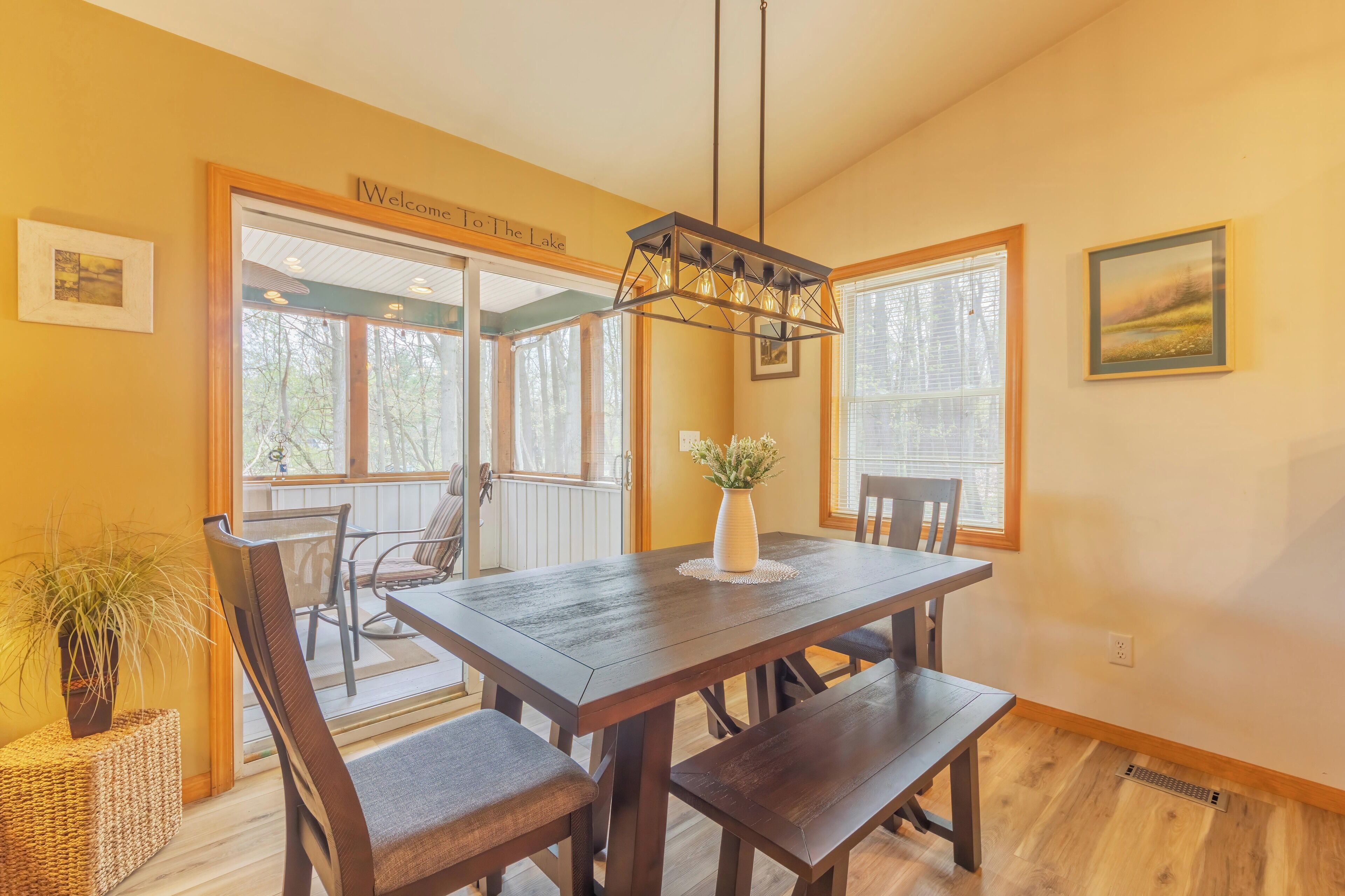 A dining area with a wooden table and chairs, a vase with flowers, and a sign reading "Welcome to the Lake".