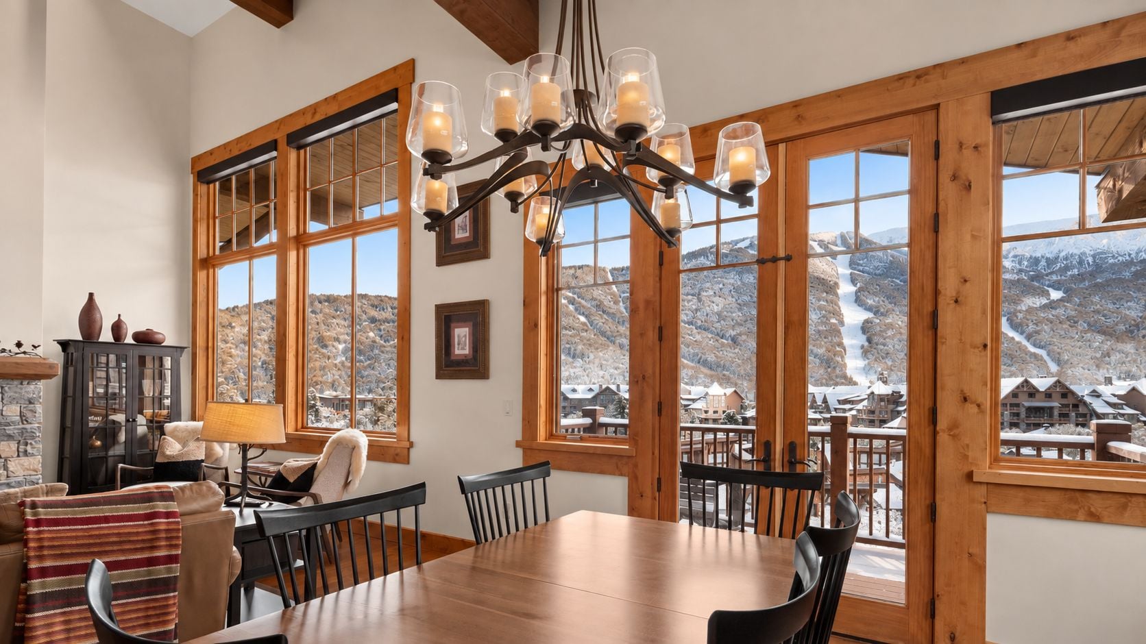 Un coin repas avec une table et des chaises en bois, de grandes fenêtres offrant une vue sur les montagnes et un lustre orné de bougies.