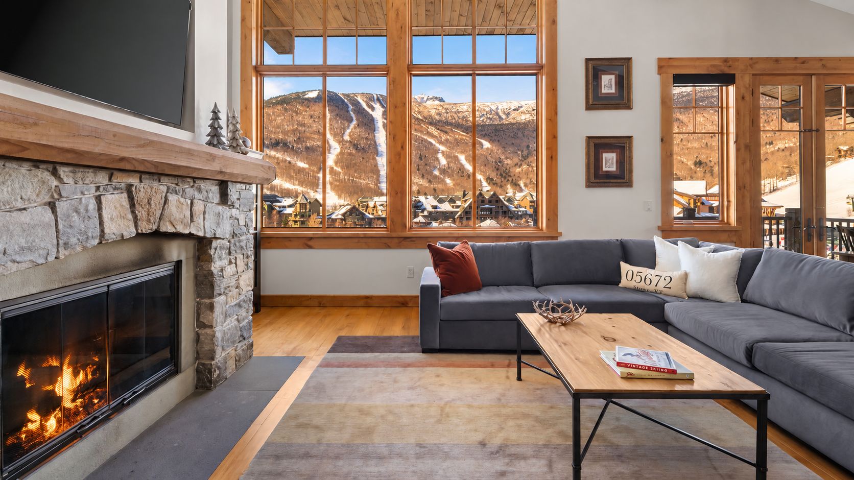 Ein Wohnzimmer mit Kamin aus Stein, einem grauen Sofa, einem Holztisch und großen Fenstern mit Bergblick.