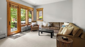 A living room with a brown sofa, a coffee table, and a chair. There are large windows with wooden frames and a view of greenery outside.