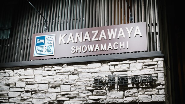 Een bordje voor Kanazawaya Showamachi met een stenen muur op de achtergrond.