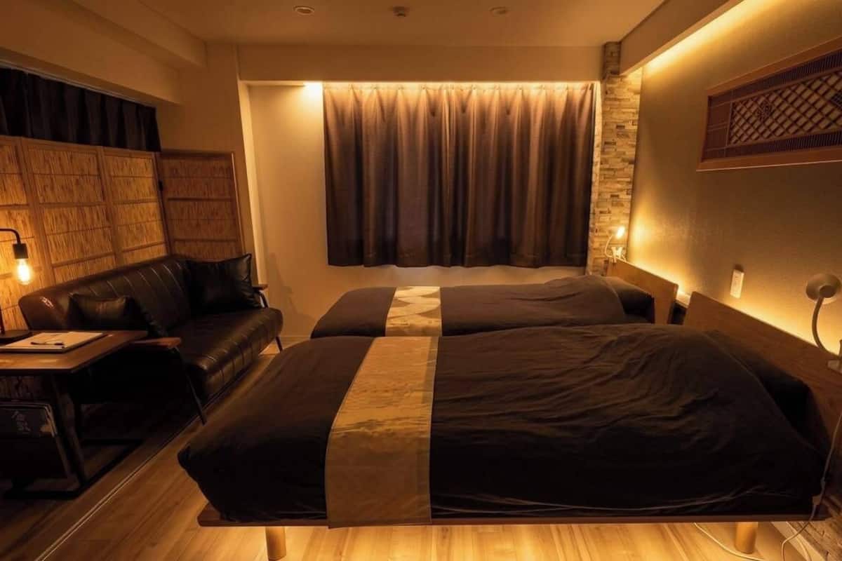 Une chambre d’hôtel avec une tête de lit en bois, un canapé, deux lits avec une literie sombre, une table de chevet et une lampe fixée au mur.
