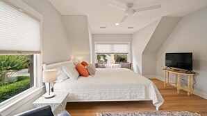 A bedroom with a large bed, a ceiling fan, a TV on a wooden table, and a view of greenery outside.