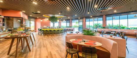 A spacious restaurant with wooden flooring, a central island with seating, and large windows offering a view of greenery.