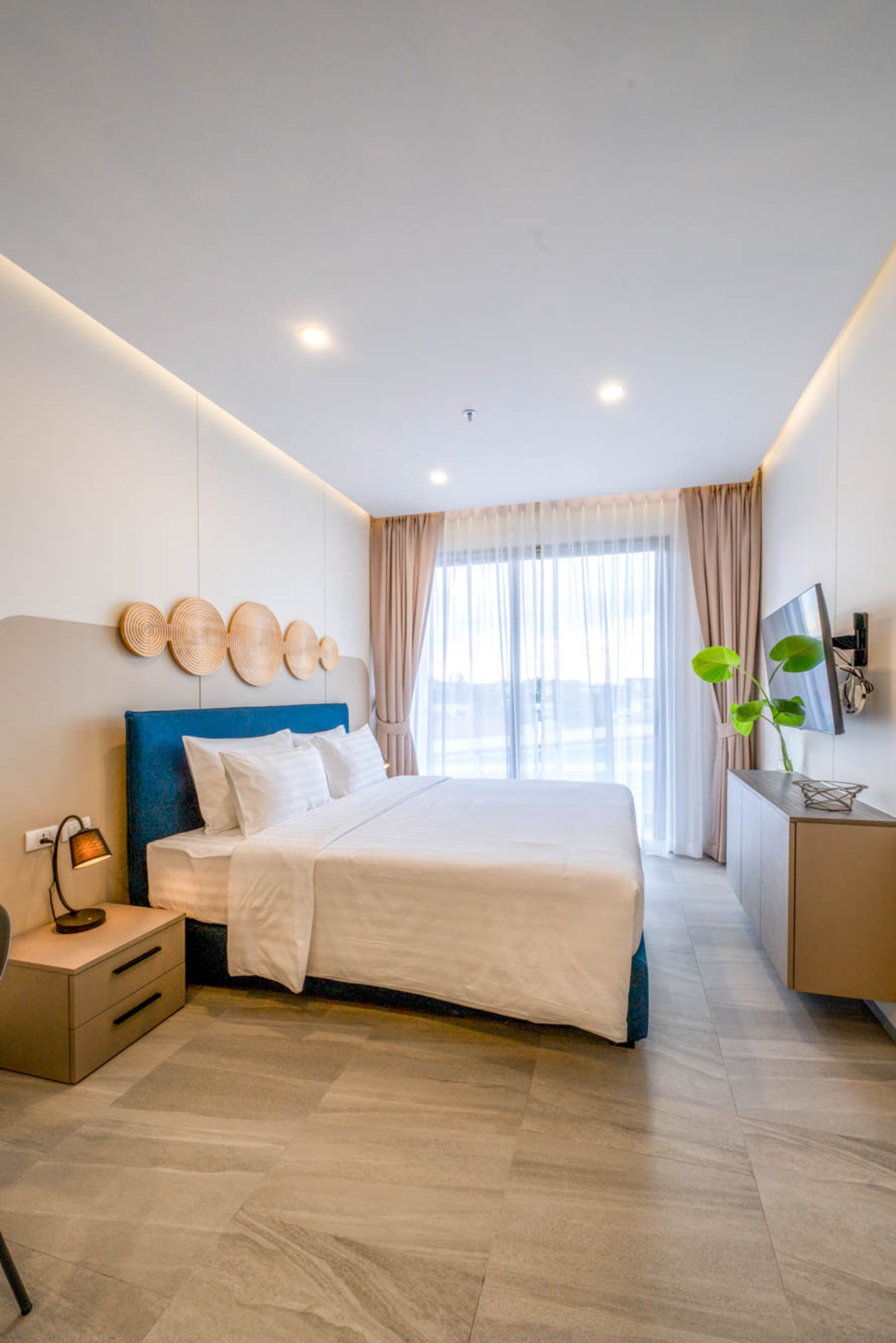 Una habitación de hotel moderna con una cama grande, una mesita de noche, un televisor montado en la pared y una ventana con cortinas.
