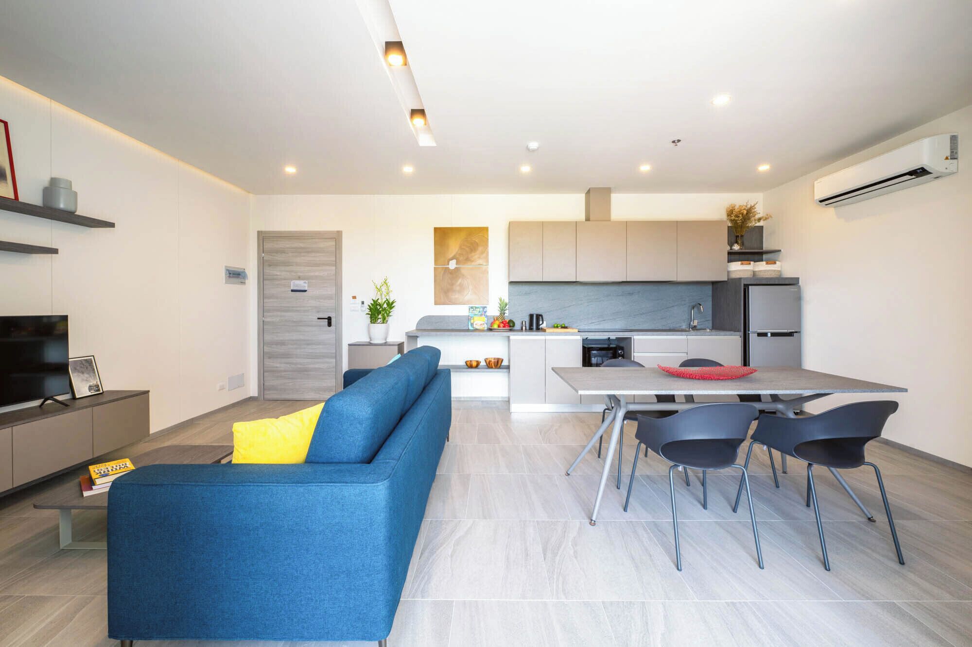 Una sala de estar moderna con un sofá azul, una mesa de comedor con cuatro sillas y una zona de cocina con refrigerador y estufa.