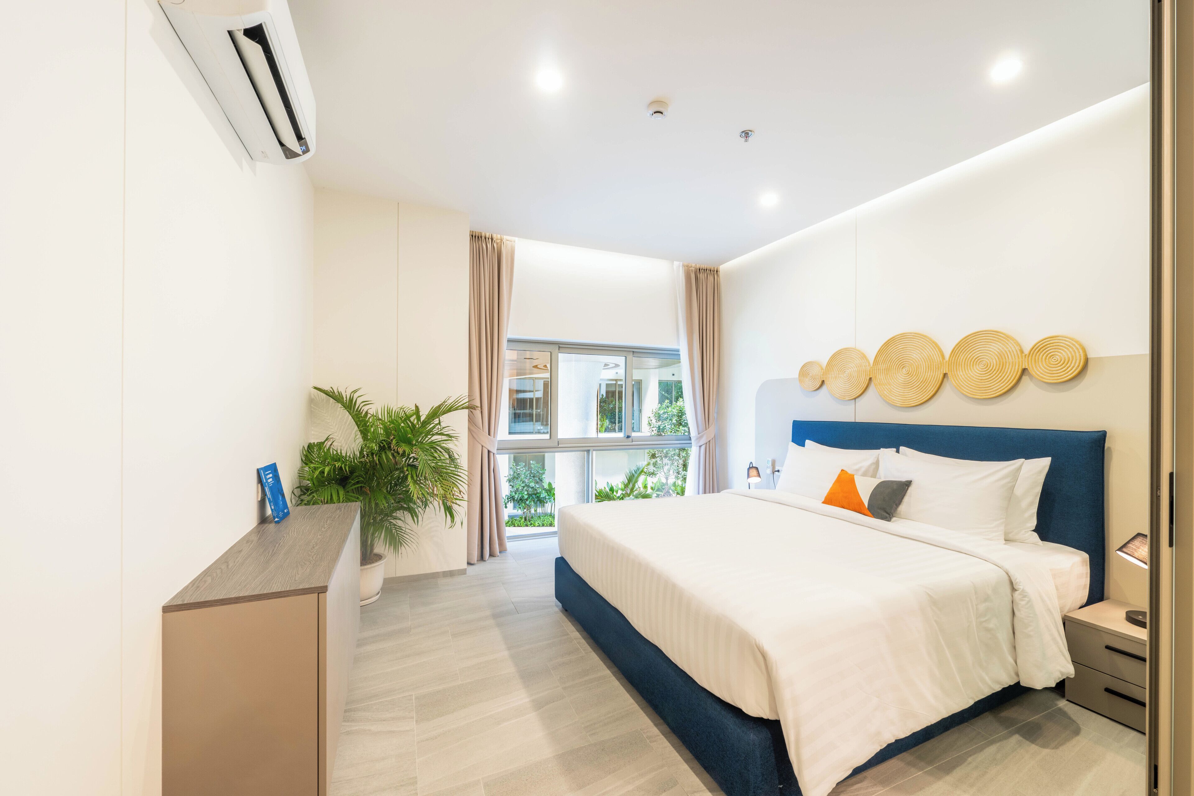 A modern hotel room with a large bed, a bedside table, a nightstand, a potted plant, and a view of the outdoors.