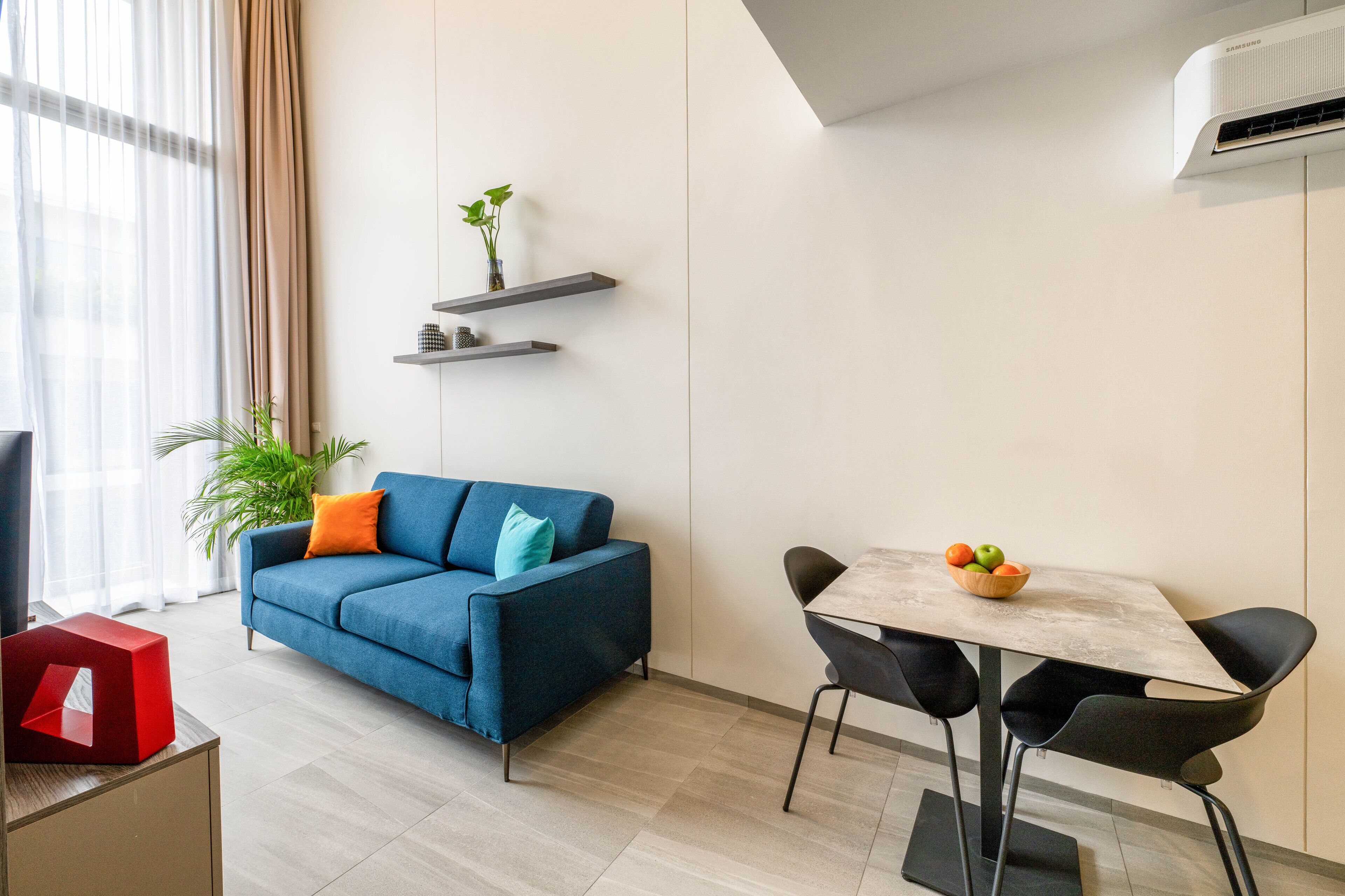Una sala de estar moderna con un sofá azul, una mesa de comedor con sillas y una pequeña planta en un estante.