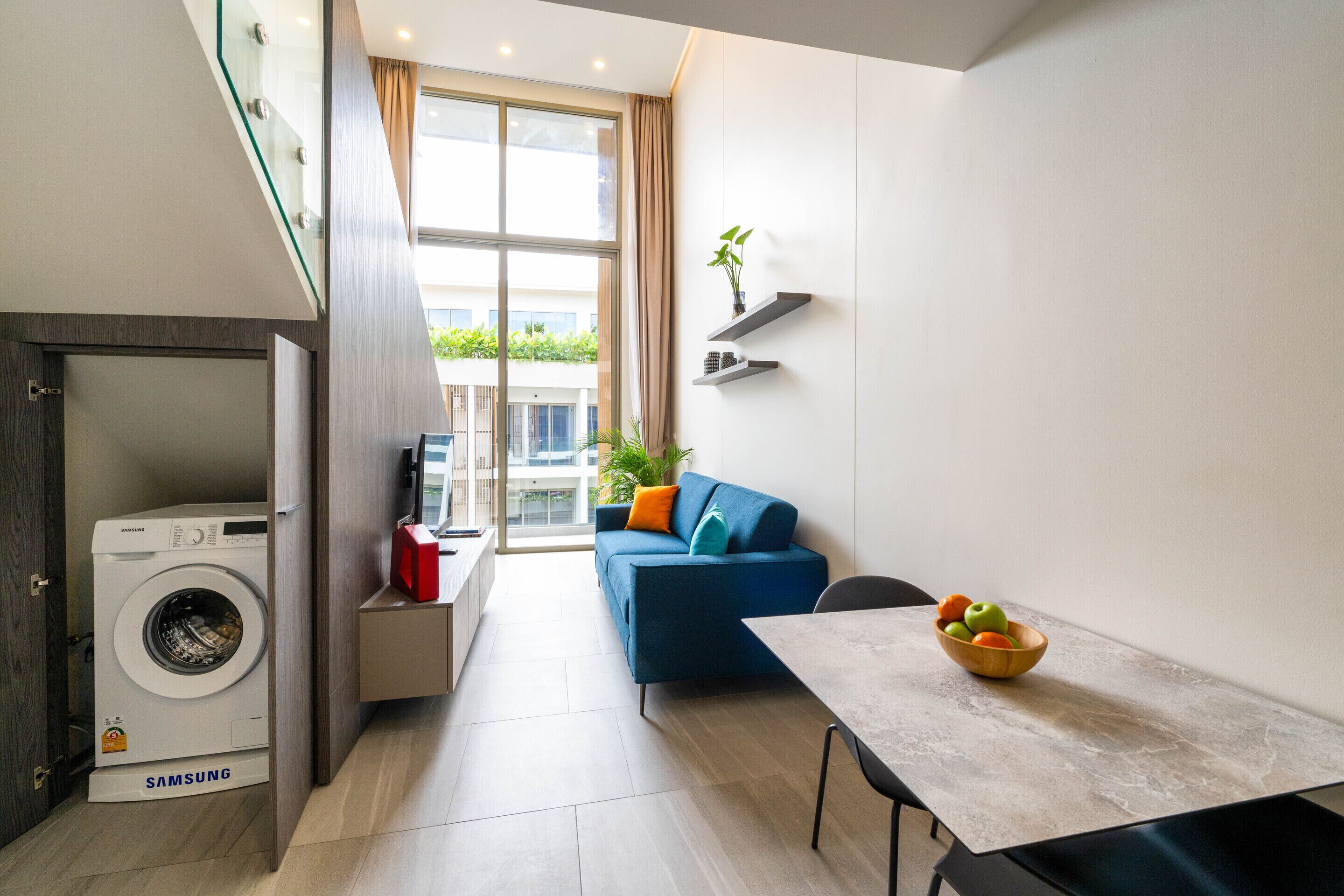 A compact living space with a washing machine, a blue sofa, a dining table, and a bowl of fruit.