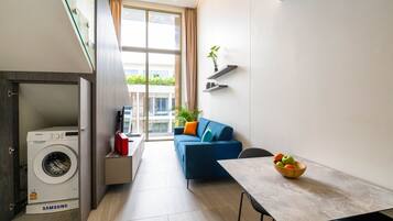 A compact living space with a washing machine, a blue sofa, a dining table, and a bowl of fruit.