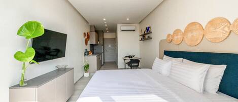 A modern hotel room with a large bed, a TV mounted on the wall, a small kitchen area, and a dining table.