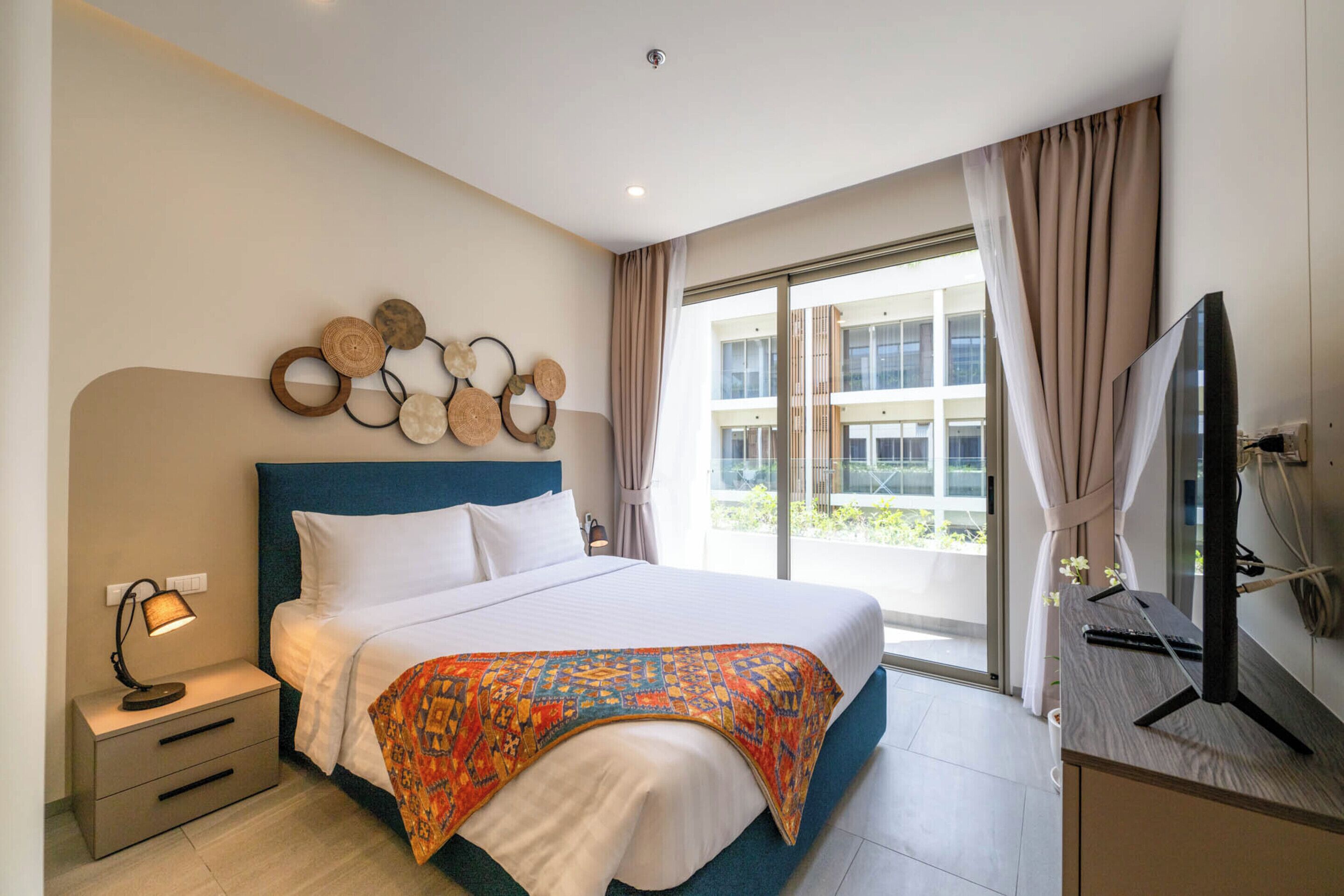 Een hotelkamer met een groot bed, een nachtkastje met een lamp, een televisie en uitzicht door een groot raam.