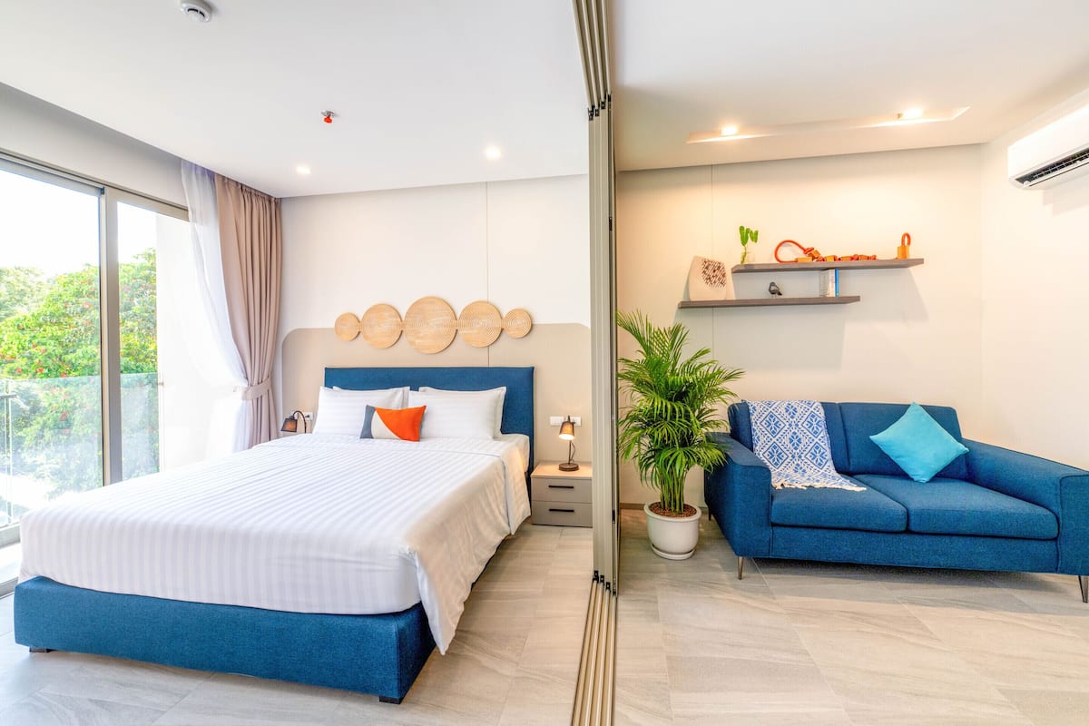 Une chambre moderne avec un grand lit, un canapé bleu, une plante en pot et une étagère murale agrémentée d’objets décoratifs.