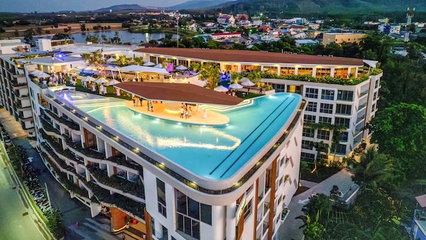 A hotel with a rooftop pool, multiple balconies, and a view of the surrounding town and hills. - HOMA Cherngtalay (Choeng Thale)