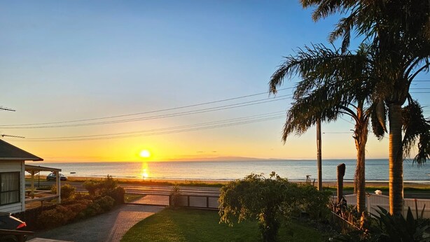 Une vue sur le coucher de soleil depuis une propriété, avec un chemin pavé menant à un portail, des palmiers et une vue sur l’océan.