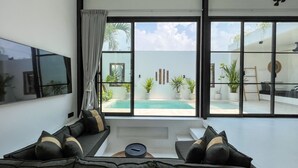A modern living room with a large TV, comfortable seating, and a view of a pool and outdoor area. - KAKAO Villas Kedungu (Tabanan)