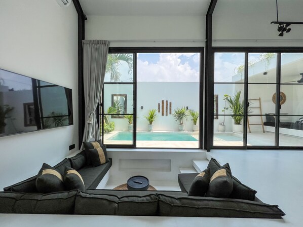 A modern living room with a large TV, comfortable seating, and a view of a pool and outdoor area.