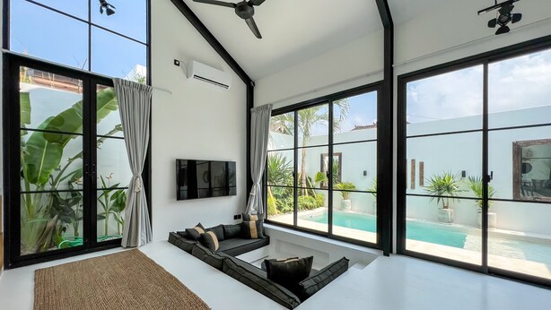 A spacious living room with a large window overlooking a pool, a ceiling fan, and a flat-screen TV mounted on the wall. - KAKAO Villas Kedungu (Tabanan)