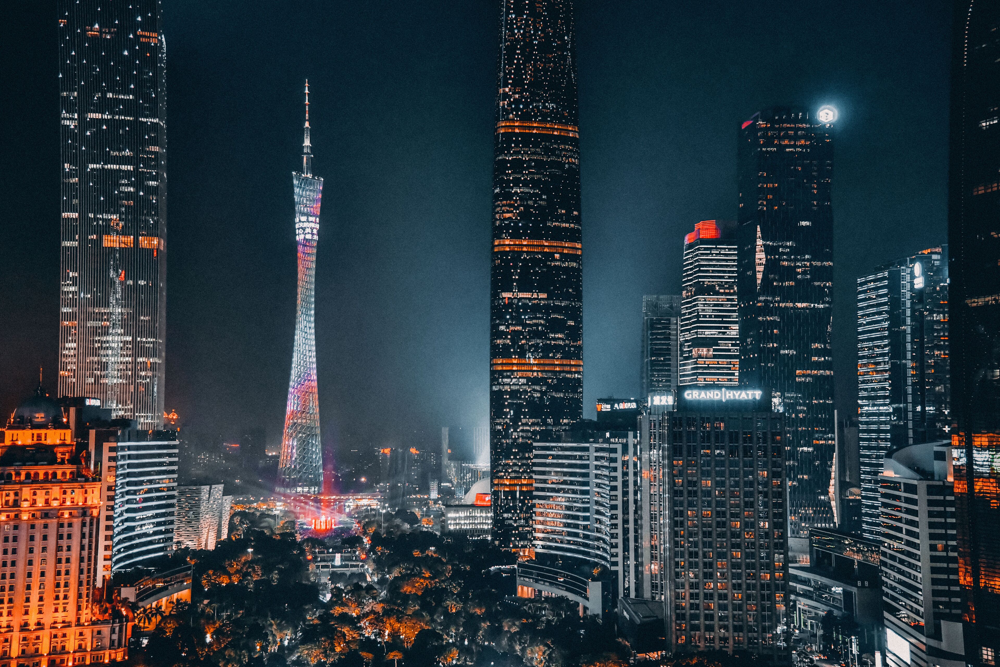 A cityscape at night with illuminated skyscrapers, including a prominent tower with a unique design and a building with a visible "Grand Hyatt" sign.