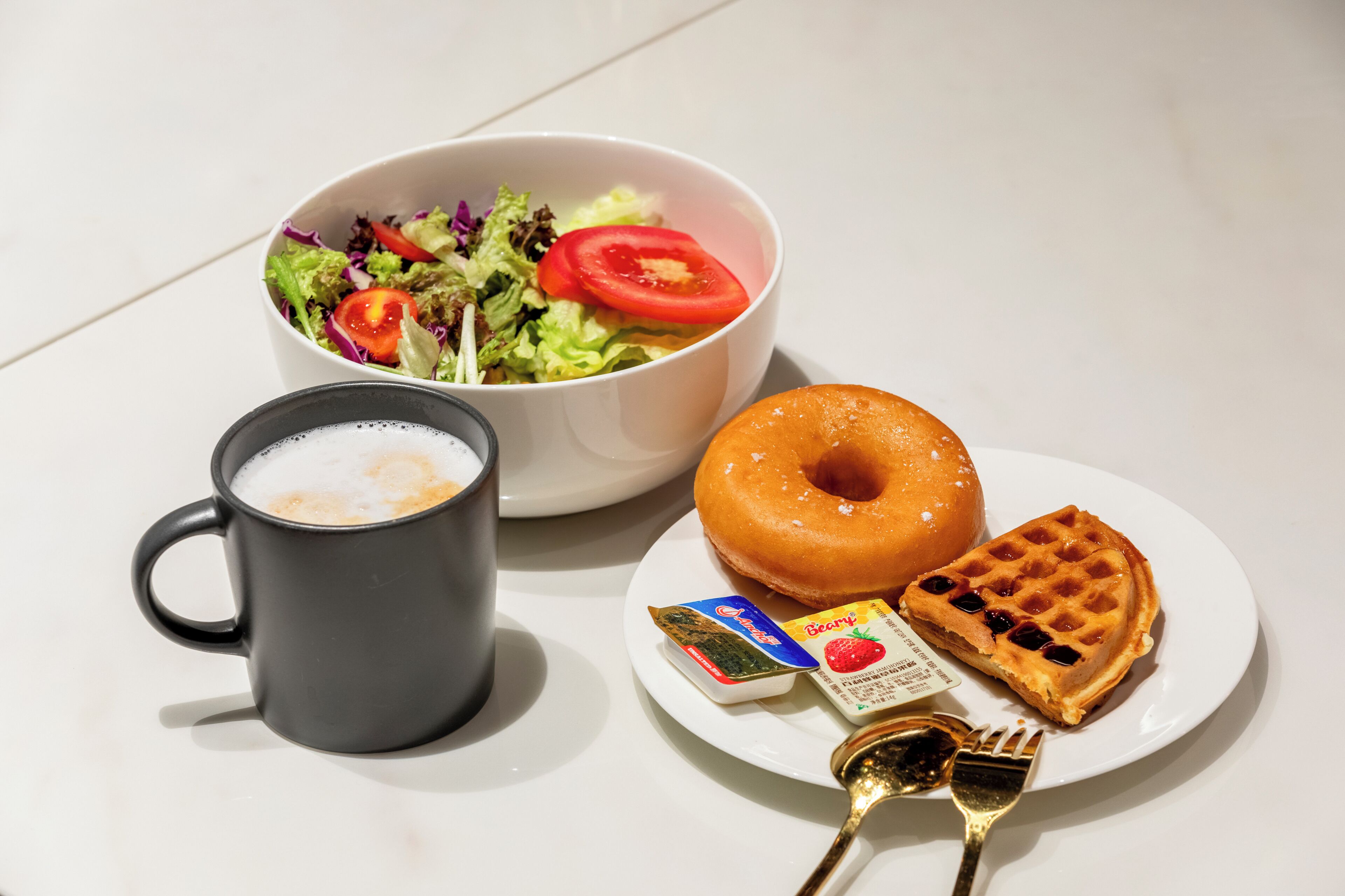 A white table with a coffee mug, a bowl of salad, a donut, a waffle, and a plate with condiments.