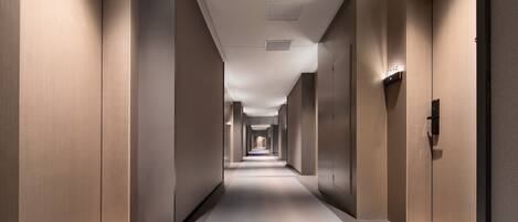 A modern hotel corridor with a straight path, light-colored walls, and a carpeted floor.